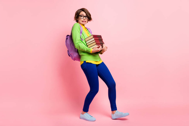 Full length photo of guilty young lady dressed yellow shirt glasses backpack walking holding book stack isolated pink color background - Photo, image