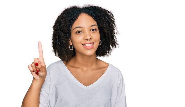 Jeune fille afro-américaine portant des vêtements décontractés montrant et pointant vers le haut avec le doigt numéro un tout en souriant confiant et heureux.  - Photo, image