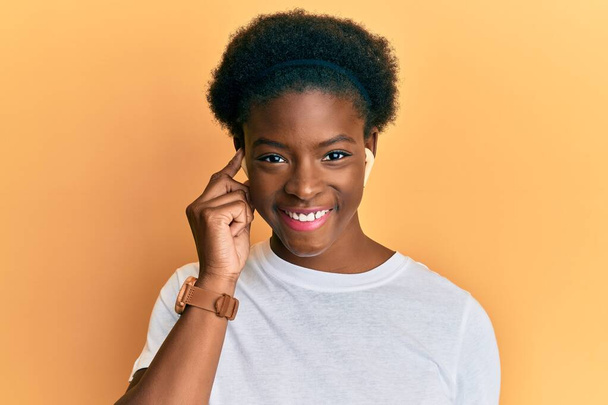 Nuori afrikkalainen amerikkalainen tyttö kuuntelee musiikkia käyttämällä kuulokkeet etsivät positiivista ja onnellinen seisoo ja hymyilee luottavainen hymy osoittaa hampaita  - Valokuva, kuva