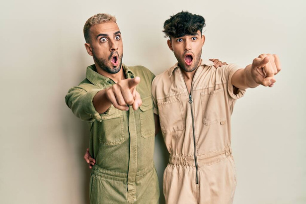 Homosexual gay couple standing together wearing casual jumpsuit pointing with finger surprised ahead, open mouth amazed expression, something on the front  - 写真・画像