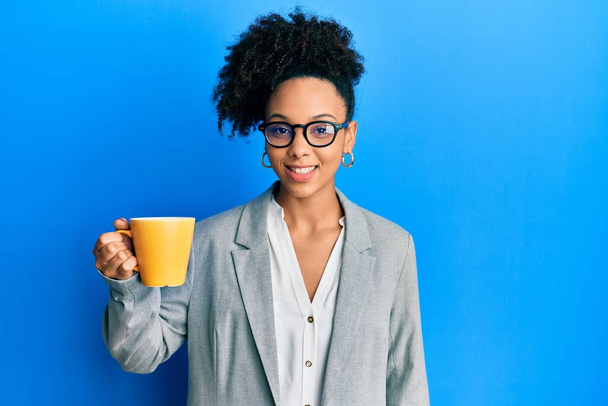 若いアフリカ系アメリカ人の少女は、ビジネス服を着てコーヒーを飲みながら、前向きな笑顔で立って笑顔で歯を見せています  - 写真・画像