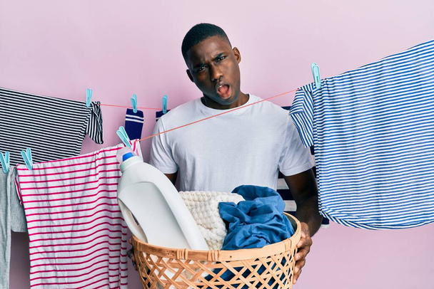 Junger afrikanisch-amerikanischer Mann hält Wäschekorb im Schockgesicht, sieht skeptisch und sarkastisch aus, überrascht mit offenem Mund  - Foto, Bild
