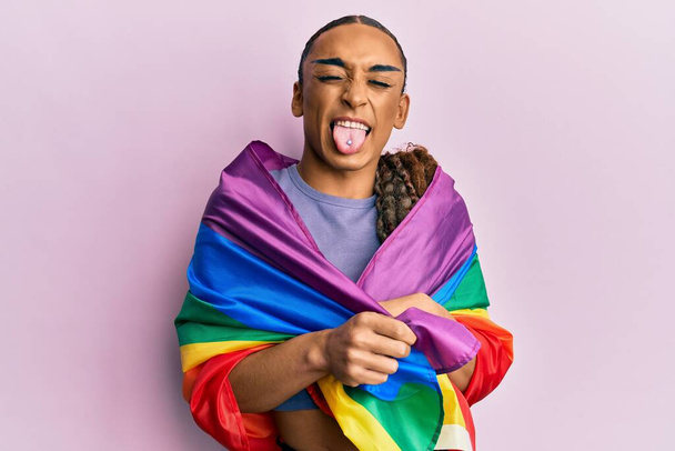 Hombre hispano usando maquillaje y pelo largo envuelto en arco iris lgbtq bandera palanca lengua hacia fuera feliz con expresión divertida.  - Foto, imagen