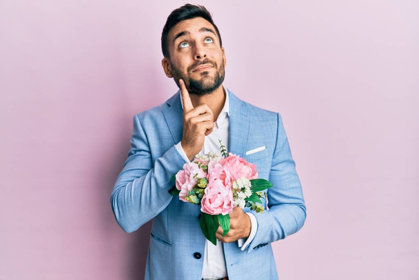 Mladý hispánský obchodník v obchodní bundě držící květiny vážný obličej přemýšlející o otázce s rukou na bradě, přemýšlivý o matoucí nápad  - Fotografie, Obrázek