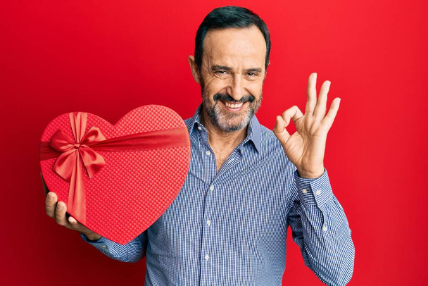 Hombre hispano de mediana edad sosteniendo regalo de San Valentín haciendo signo de ok con los dedos, sonriendo gesto amistoso excelente símbolo  - Foto, Imagen