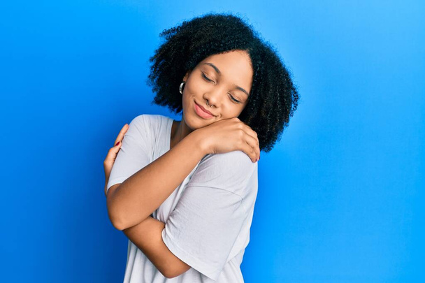 Jovem afro-americana vestindo camisa branca casual abraçando-se feliz e positiva, sorrindo confiante. auto-amor e auto-cuidado  - Foto, Imagem