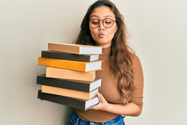 Jovencita hispana sosteniendo un montón de libros haciendo cara de pez con boca y ojos entrecerrados, loca y cómica.  - Foto, Imagen