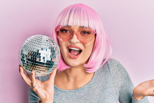 Joven mujer caucásica vistiendo peluca rosa sosteniendo bola disco celebrando logro con sonrisa feliz y expresión ganadora con la mano levantada  - Foto, Imagen