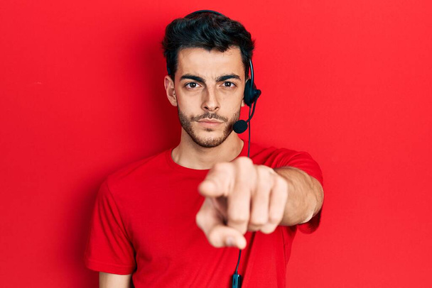 Νεαρός Ισπανός που φοράει ακουστικά τηλεφωνικού κέντρου δείχνοντας με το δάχτυλο την κάμερα και σε σένα, με αυτοπεποίθηση δείχνει σοβαρός.  - Φωτογραφία, εικόνα