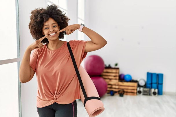 African american woman with afro hair holding yoga mat at pilates room smiling cheerful showing and pointing with fingers teeth and mouth. dental health concept.  - Zdjęcie, obraz