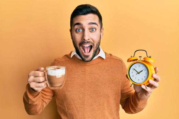 Young hispanic man holding coffee and alarm clock celebrating crazy and amazed for success with open eyes screaming excited.  - Photo, image