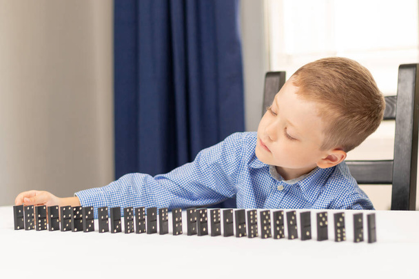 Ein sechsjähriger süßer Junge spielt zu Hause auf einem weißen Holztisch Dominosteine. Selektiver Fokus. Nahaufnahme - Foto, Bild
