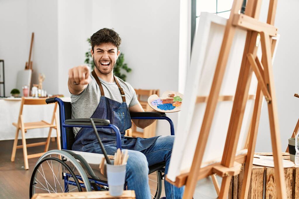 Jeune homme hispanique assis sur la peinture en fauteuil roulant au studio d'art pointant mécontent et frustré à la caméra, en colère et furieux contre vous  - Photo, image
