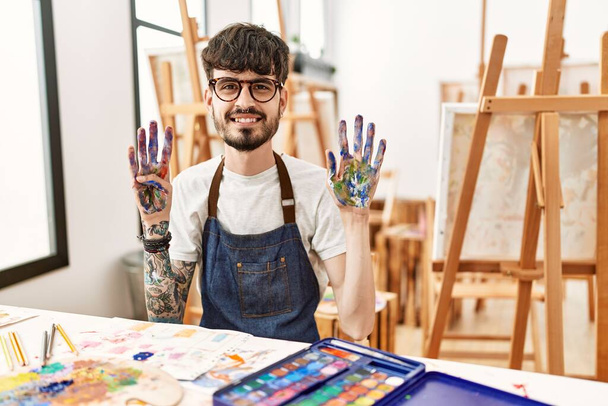 Spanyol férfi szakállal a művészeti stúdióban bemutatja és felmutat a kilences számú ujjaival, miközben magabiztosan és boldogan mosolyog..  - Fotó, kép