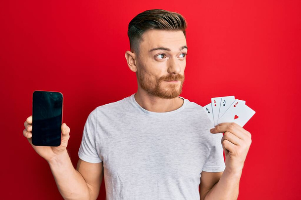 スマートフォンとカジノのカードを持っている若い赤毛の男は、横を見て笑って離れて考えて見つめていた.  - 写真・画像