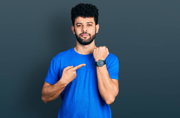 Молодой араб с бородой в повседневной синей футболке в спешке указывает на часы, нетерпение, смотрит в камеру с расслабленным выражением лица  - Фото, изображение