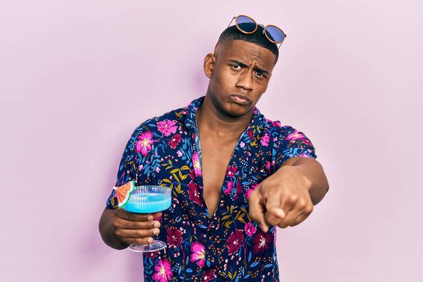 Genç siyahi adam alkolle tropikal kokteyl içiyor. Kamerayı işaret ediyor. Kendine güveni tam bir jest.  - Fotoğraf, Görsel