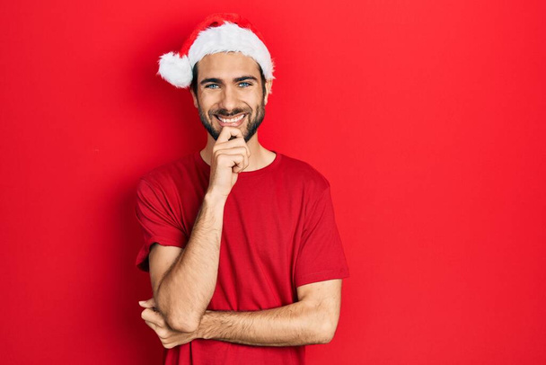 Young hispanic man wearing christmas hat looking confident at the camera with smile with crossed arms and hand raised on chin. thinking positive.  - Valokuva, kuva