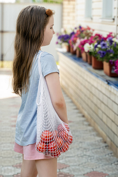Na dvoře domu stojí malá holčička s bílým síťovaným pytlíkem s rajčaty přes rameno. Moderní opakovaně použitelné nákupy a koncepce nulového odpadu. - Fotografie, Obrázek