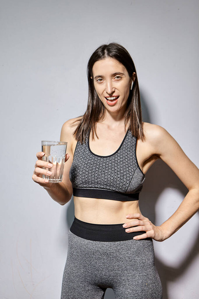 Una mujer en ropa deportiva y con auriculares inalámbricos bebe agua. el concepto de un estilo de vida saludable y actividades deportivas - Foto, imagen