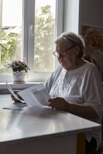 Starsza kobieta siedząca przy stole w kuchni w domu trzyma krajowe rachunki, czuje się zaniepokojona, że zapomniała zapłacić lub powstał dług. Emerytka w średnim wieku - Zdjęcie, obraz