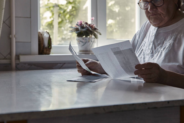 Elderly woman sitting at table in kitchen at home holds domestic bills, feels concerned forgot to pay or debt formed. Middle aged 80s retired woman - Photo, Image