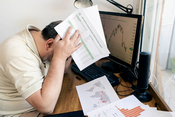 A Caucasian man holding his head worried about global economic losses with papers with printed calculation tables in his hands and on the desk. concept globalization, economy, frustration, anger, concern. - Photo, Image