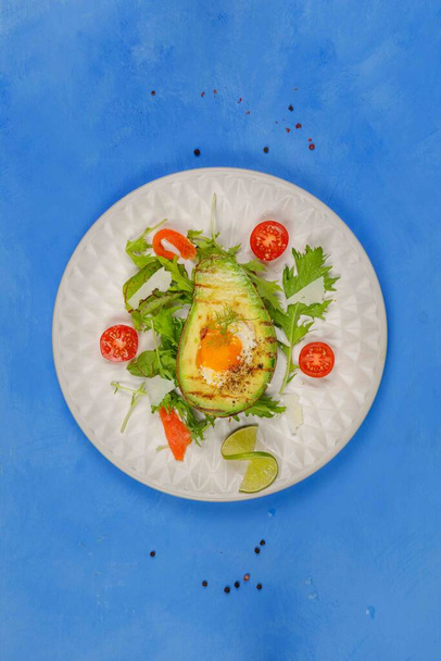 Oryginalne smażone jajka pieczone w awokado, podawane z liśćmi sałatki, pomidorami i parmezanem. Zdrowe, pożywne danie o wysokiej zawartości białka, superjedzenie. Jaja smażone z awokadą.Pionowa rama, góra, na niebieskim tle, kopia przestrzeni - Zdjęcie, obraz