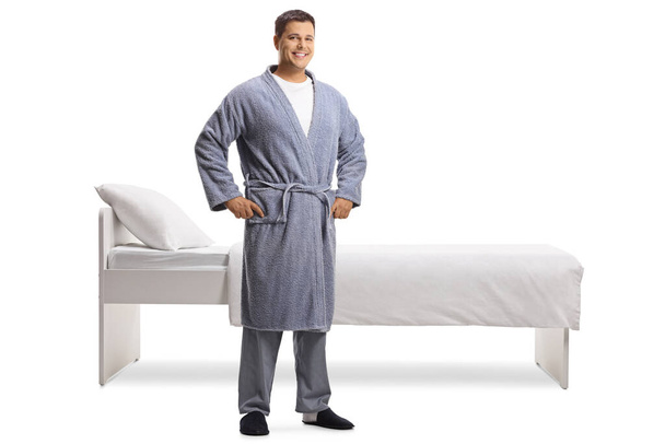 Full length portrait of a young man wearing a robe and standing in front of a bed isolated on white background - Photo, image