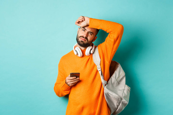 Tired guy with headphones and backpack, wiping sweat off forehead with exhausted face, using mobile phone, standing over turquoise background - Photo, Image