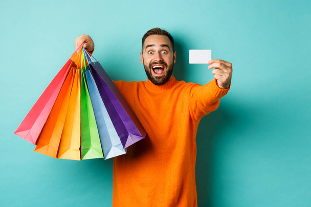 Glücklicher Mann mit Kreditkarte und Einkaufstaschen vor türkisfarbenem Hintergrund - Foto, Bild