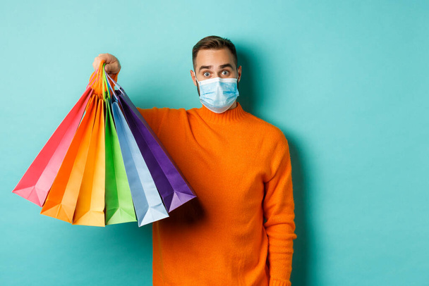 Covid-19, social distancing and lifestyle concept. Young man in face mask showing shopping bags, buying holiday gifts during pandemic, standing over blue background - Foto, imagen
