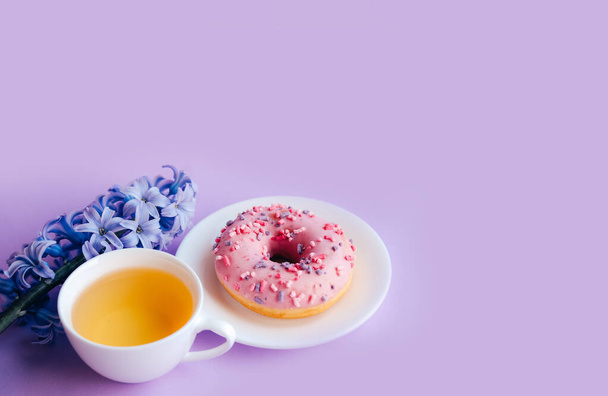 Roze suiker geglazuurde donut, kopje thee en paarse hyacint bloem op kleurrijke achtergrond. - Foto, afbeelding