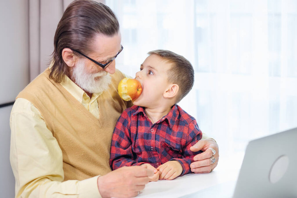 Komea isoisä ja pojanpoika käyttävät vempaimia ja hymyilevät viettäessään aikaa yhdessä kotona. Pojanpoika leikkii omenalla. - Valokuva, kuva