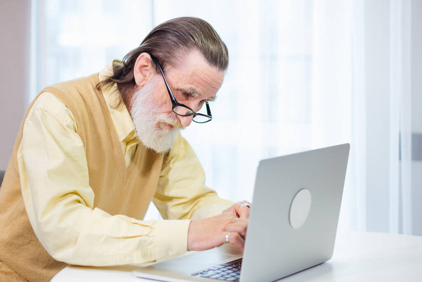 Dziadek uczy się pracować na laptopie. Rachunki są płacone w domu w kuchni. Technologie internetowe dla osób starszych. Emerytowany starzec pracujący nad komputerowym uczeniem się w sieciach społecznościowych - Zdjęcie, obraz