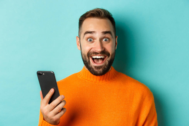 Zbliżenie szczęśliwego przystojniaka piszącego wiadomość przez telefon komórkowy, trzymającego smartfona i uśmiechającego się, stojącego na turkusowym tle - Zdjęcie, obraz