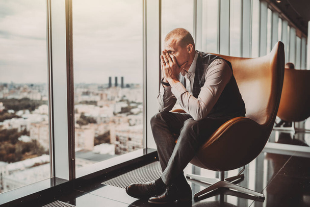 A stressed out adult caucasian man entrepreneur partly closing face with his hands while sitting on an office armchair next to the window of a modern business skyscraper and looking on the cityscape - Foto, immagini