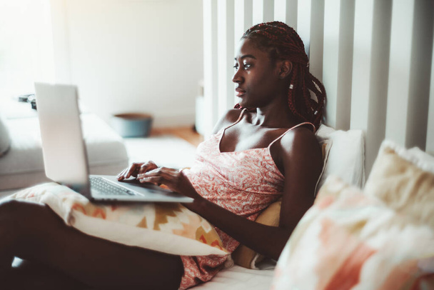 Чуттєва красива чорна жінка з червоним плетеним волоссям і в нічному одязі лежить на ліжку в приміщенні її квартири вранці і використовує ноутбук для перегляду фільмів і спілкування з друзями
 - Фото, зображення