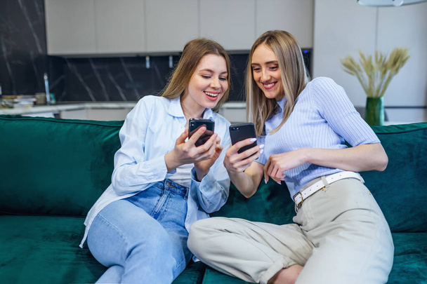 подружки сидят дома на диване, показывая друг другу фотографии отпуска на современном мобильном телефоне - Фото, изображение