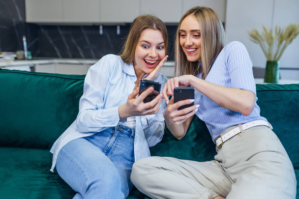 Young female students, smiling, happy sitting on the couch in the living room, showing something to each other on a mobile phone. The girl is holding a smartphone, she is in shock, one won a coupon for a purchase on the Internet - Photo, Image