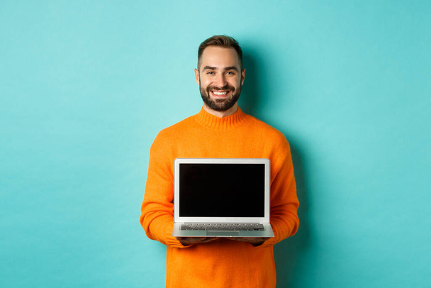 Jóképű szakállas férfi narancssárga pulóverben mutatja laptop képernyőn, bemutató online áruház, álló felett világoskék háttér - Fotó, kép