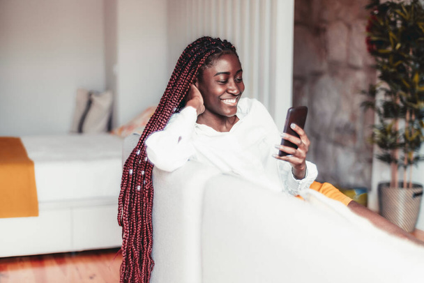 Um retrato de uma jovem mulher negra alegre com o cabelo trançado ter uma chamada de vídeo telefone ou uma transmissão on-line vlogging enquanto se senta no sofá de sua sala de estar casa e sorrindo - Foto, Imagem
