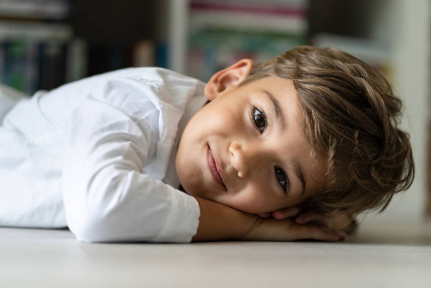 Πορτρέτο του μικρού καυκάσιου αγοριού που βρίσκεται στο πάτωμα στο σπίτι κοιτάζοντας την κάμερα με χαμόγελο στο πρόσωπό του - Φωτογραφία, εικόνα