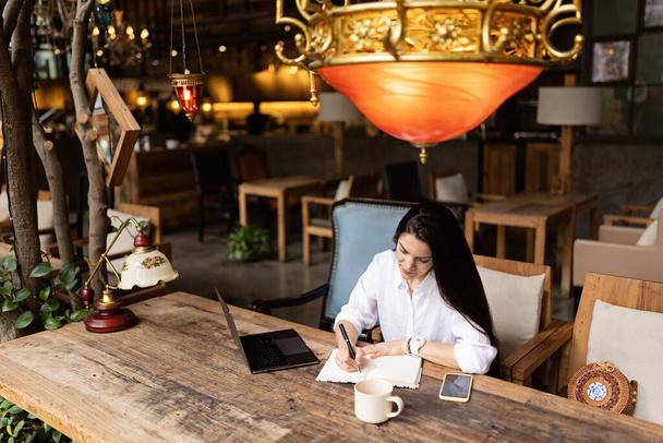 Młoda kobieta biznesu kaukaskiego z długimi brunetkami pracująca na laptopie w kawiarni. Studentka korzystająca z technologii, edukacji online, freelance  - Zdjęcie, obraz
