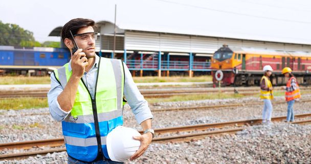 Skuteczni specjaliści ds. logistyki kolejowej lub inżynierowie kolejowi w kaskach i kamizelkach bezpieczeństwa stojący na zewnątrz toru kolejowego posiadają krótkofalówkę sprawdzającą gotowość do pracy - Zdjęcie, obraz