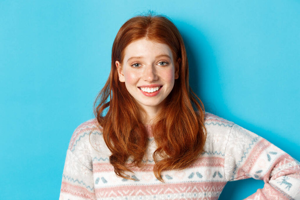 Крупный план привлекательной студентки с рыжими волосами, в зимнем свитере, улыбающейся счастливой и смотрящей в камеру, синий фон - Фото, изображение