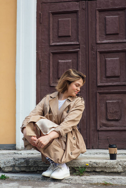 Young millennial woman with wild hair dressed in an autumn coat sitting near the door of an old building with coffee to go. - Φωτογραφία, εικόνα