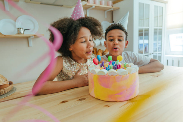Ευτυχισμένο κορίτσι κοιτάζοντας την τούρτα γενεθλίων με κεριά να διασκεδάζουν κατά τη διάρκεια του πάρτι γενεθλίων στην κουζίνα. Επιλεκτική εστίαση στο πρόσωπο του κοριτσιού. - Φωτογραφία, εικόνα