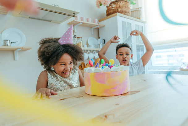 Счастливая девушка смотрит на именинный торт со свечами, весело проводя время во время празднования дня рождения на кухне. Выборочный фокус на лице девушки. - Фото, изображение