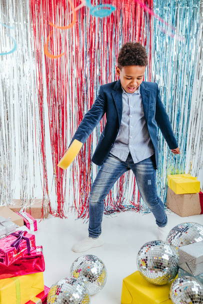 Niño feliz cantando usando piruleta decorativa en el estudio decorado con colorido fondo de cortina de papel de aluminio y bolas de discoteca y cajas de regalo. - Foto, imagen